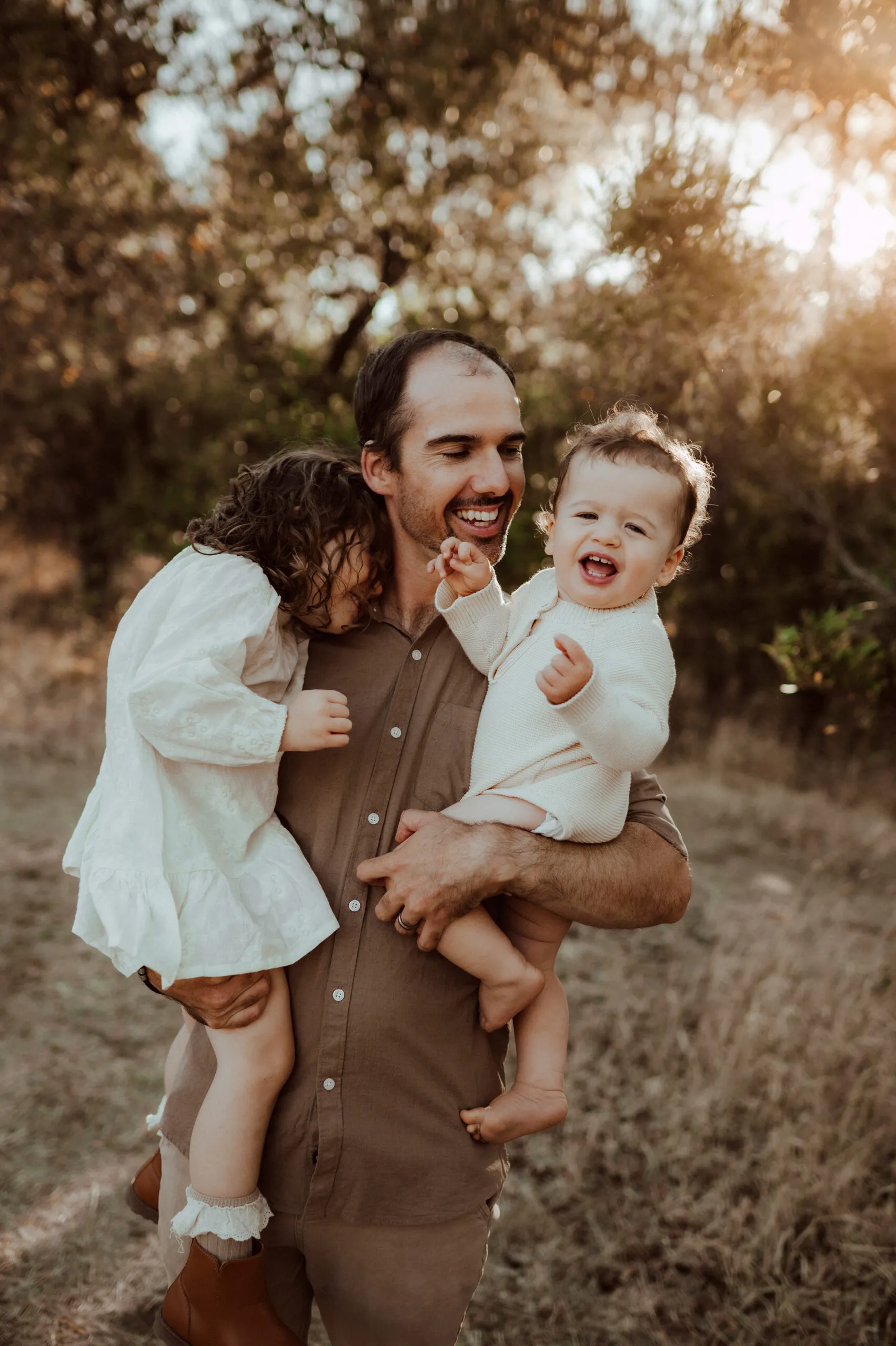 A smiling dad holding two small children outdoors in warm evening light